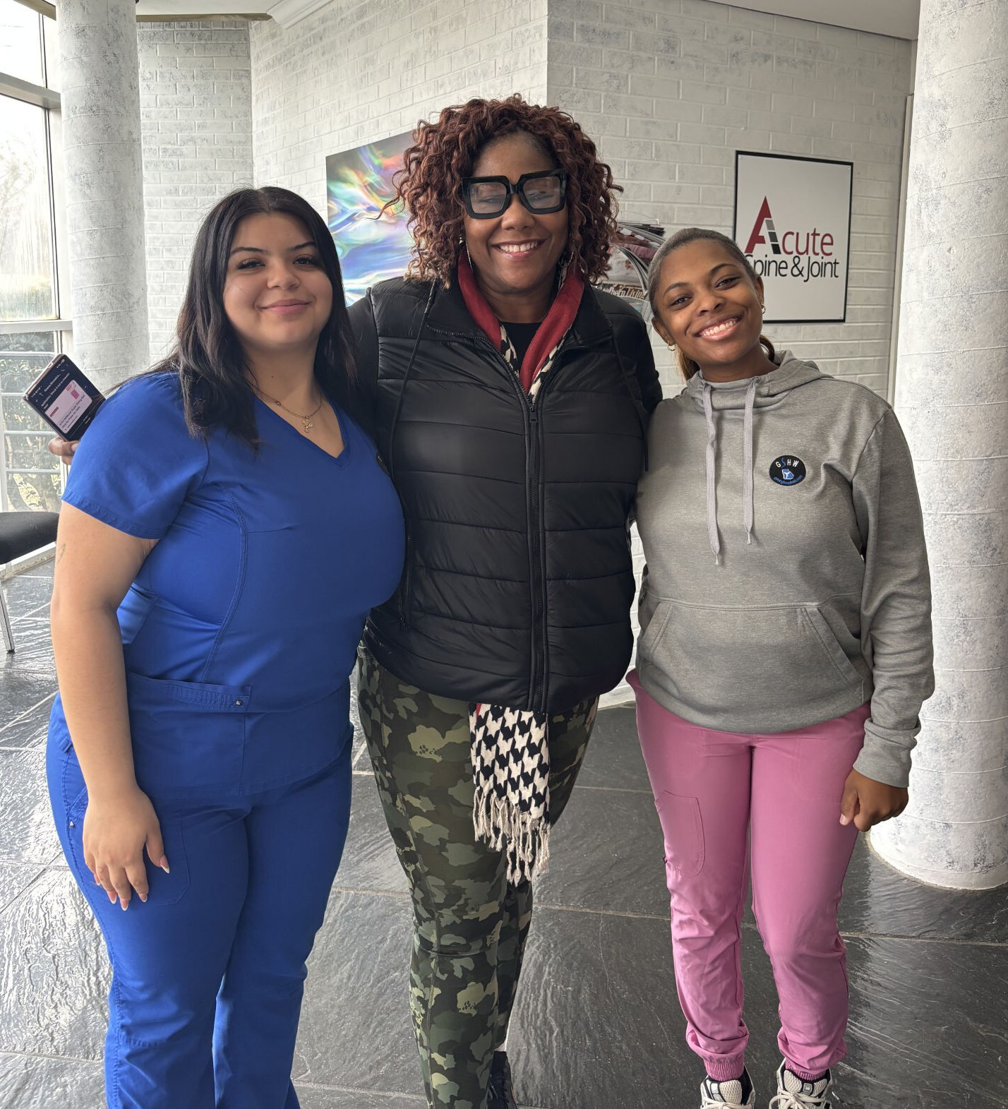 Three women stand together indoors, smiling at the camera. One wears blue scrubs, another wears a black jacket and camouflage pants, and the third wears a gray hoodie and pink pants.