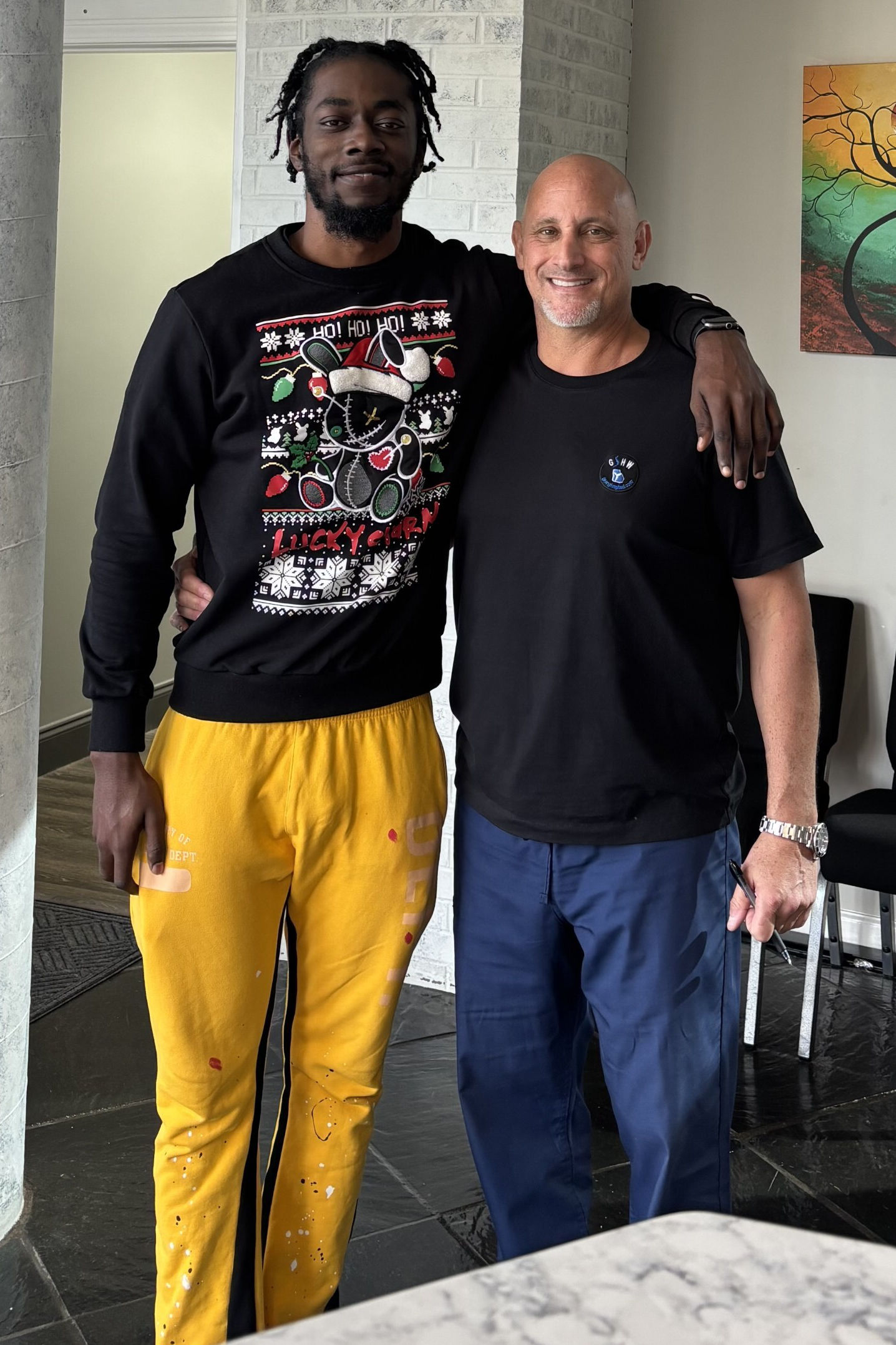 Two men stand side by side indoors, one with his arm around the other's shoulder. One wears a festive sweater and yellow pants; the other wears a black t-shirt and blue pants.