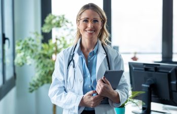 Portrait of attractive mature female doctor looking at camera while holding digital tablet standing in the consultation.