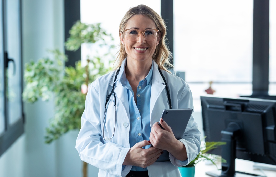 Portrait of attractive mature female doctor looking at camera while holding digital tablet standing in the consultation.