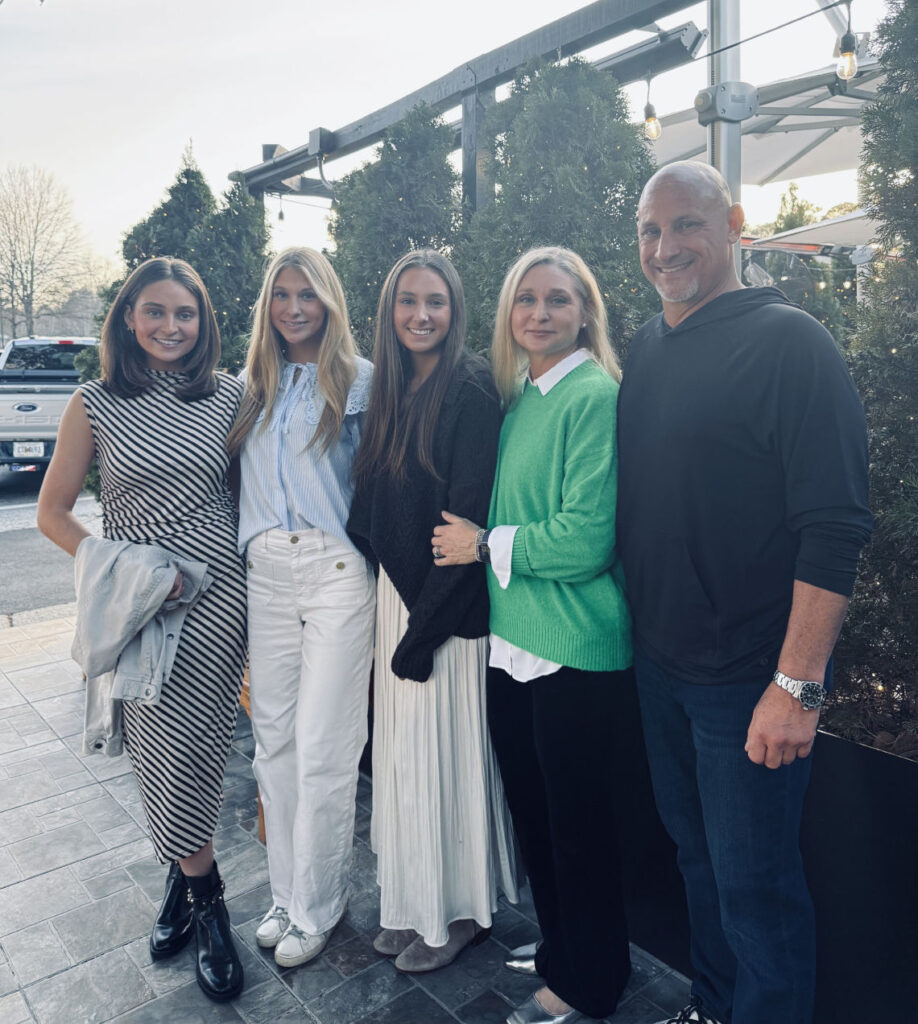 Five people stand together outdoors on a paved walkway, posing for a group photo in front of greenery and a metal railing.