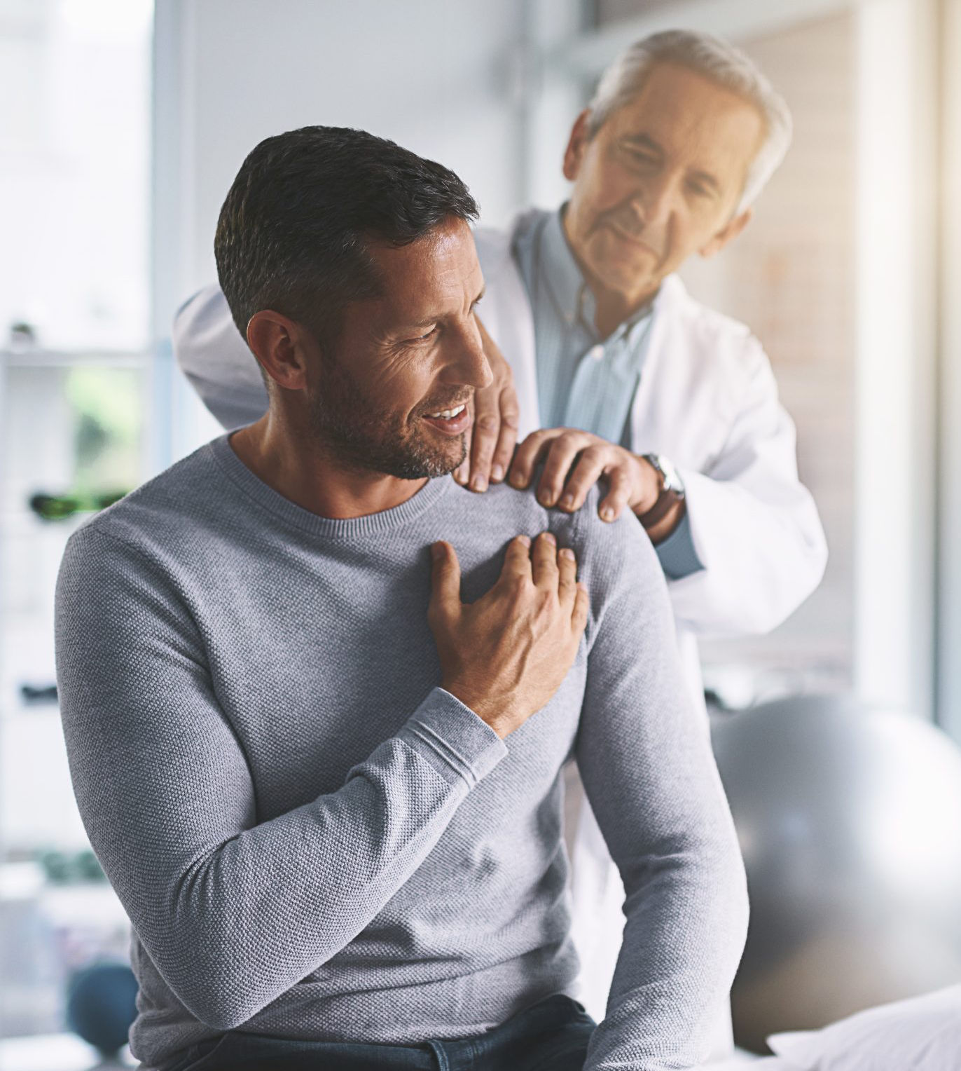 A doctor examines a man's shoulder in a medical office; the man is seated and appears to be describing his discomfort.