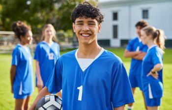 Young athlete in blue uniform holding soccer while smiling and looking at camera. Portrait of happy player ready for team training at high school. Young man during training on soccer field.