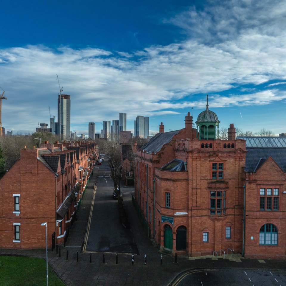 View down a narrow street lined with red-brick row houses and a large historic brick building with a green-domed tower in the foreground, leading toward a modern city skyline with tall glass skyscrapers in the distance under a partly cloudy blue sky.