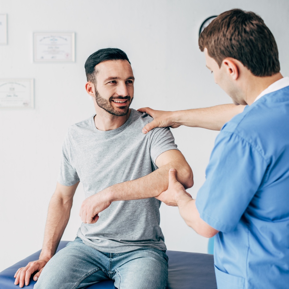 Smiling patient sitting on couch and doctor examining patient shoulder in massage cabinet at clinic