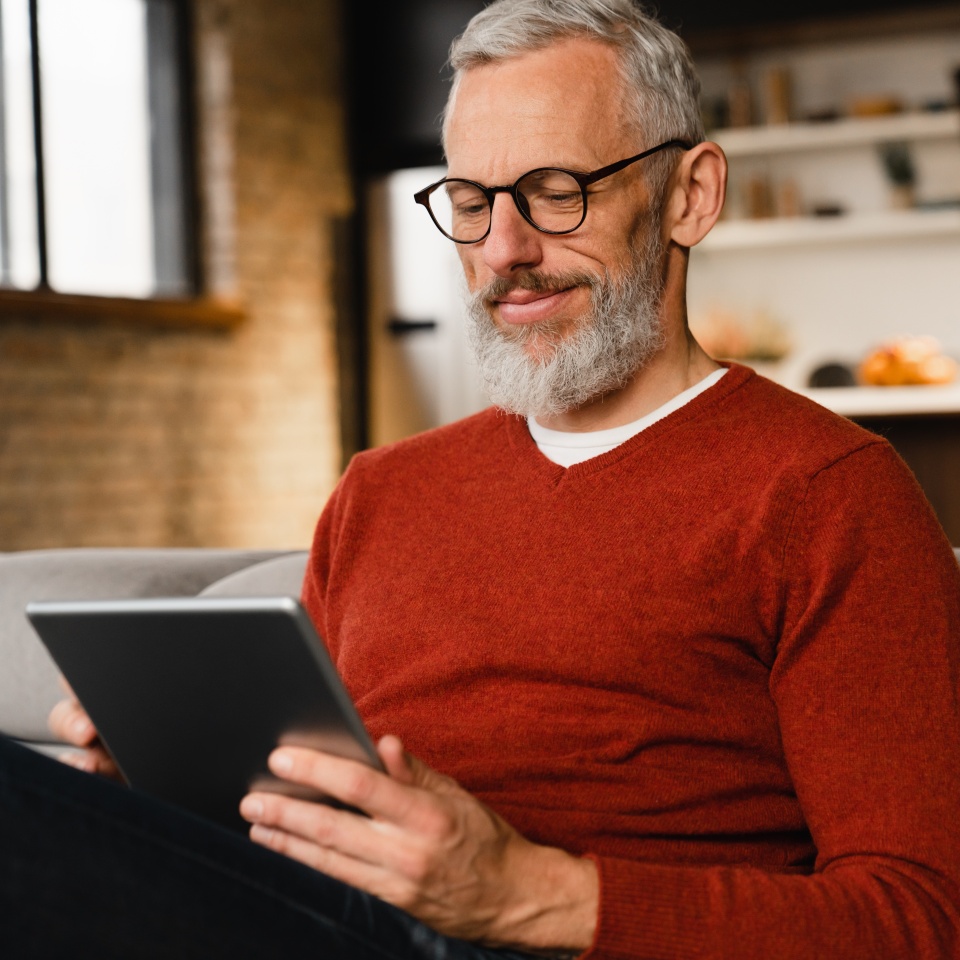 Closeup portrait of a smiling caucasian middle-aged mature man in glasses using digital tablet for e-learning, paying bills online, e-commerce, remote work, social media online at home