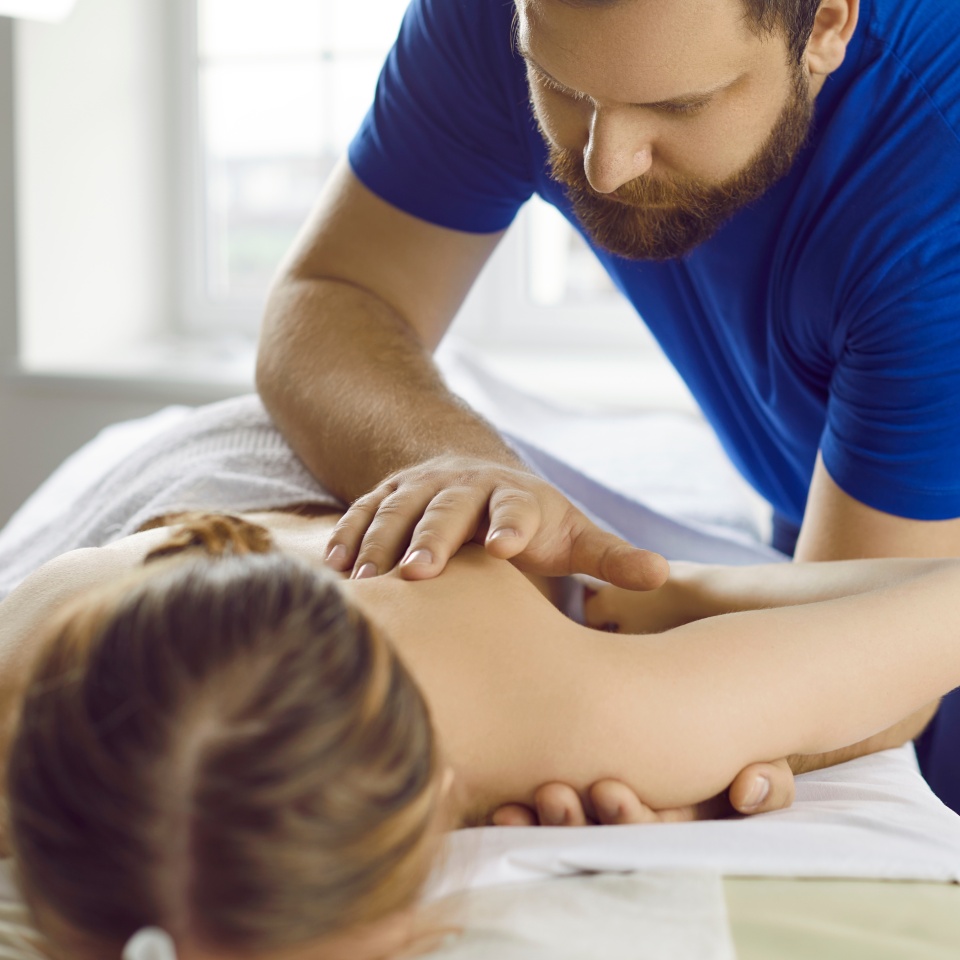 Woman is receiving treatment at physiological center. Professional male masseur therapist performs therapeutic body massage for young woman. Man massages shoulders of woman lying on massage table.