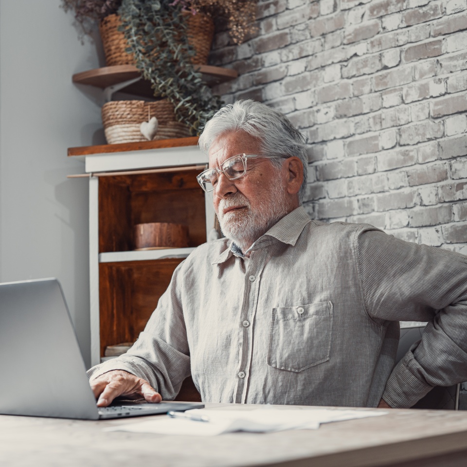 Grey haired middle aged senior male sitting alone in the kitchen on chair stretching back after long time working on computer doing exercises for loins preventing osteoarthritis and spinal stenosis