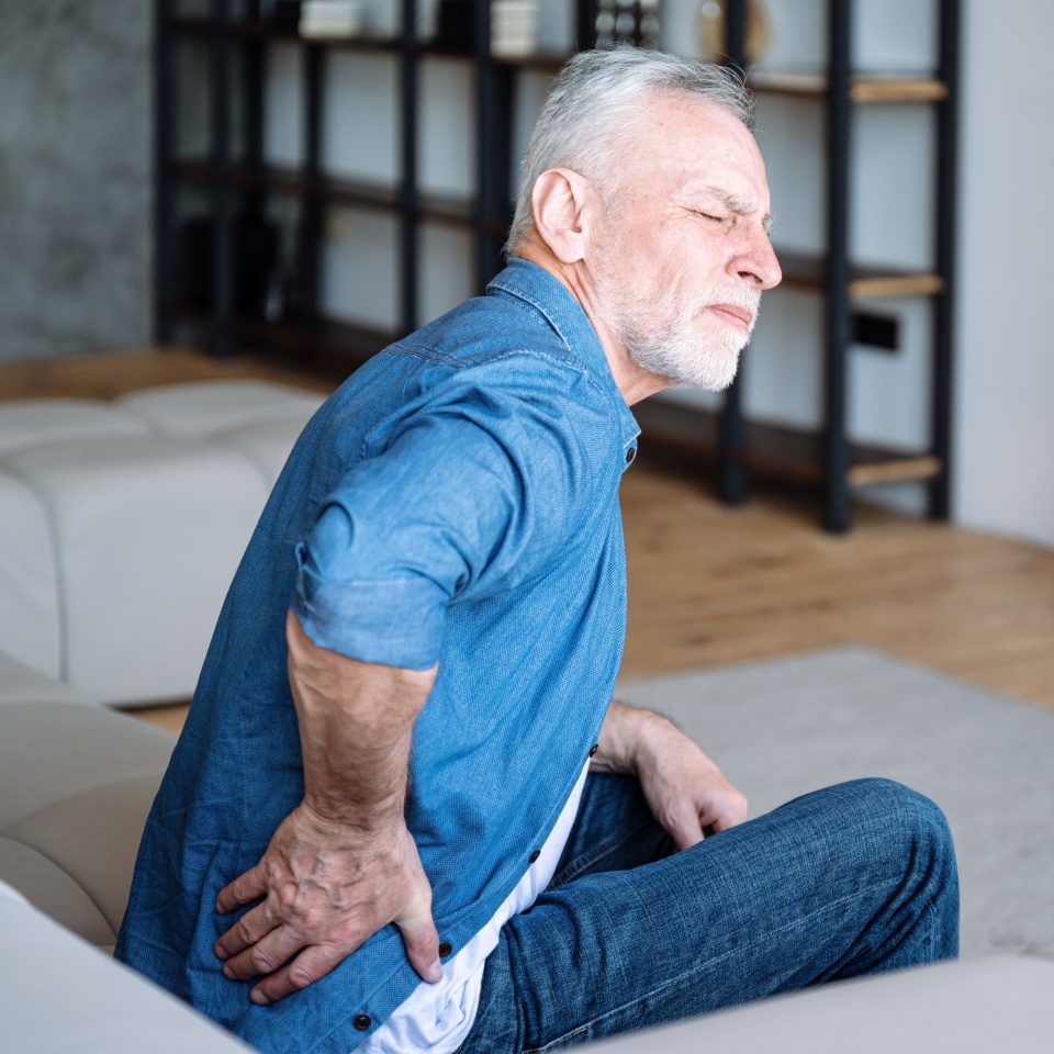 Senior man feeling pain, spasm, backache. Mature male sitting on sofa and have cramps in lower back. Suffering from chronic illness, problem with standing up. Lumbar osteochondrosis, inflammation