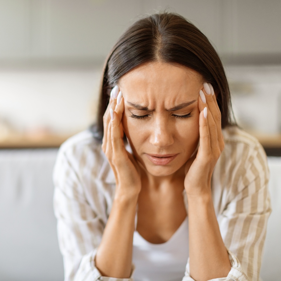 Woman in distress with severe headache, pressing her temples and frowning, young female showing grimace of pain while sitting on couch in bright living room, having signs of stress or migraine