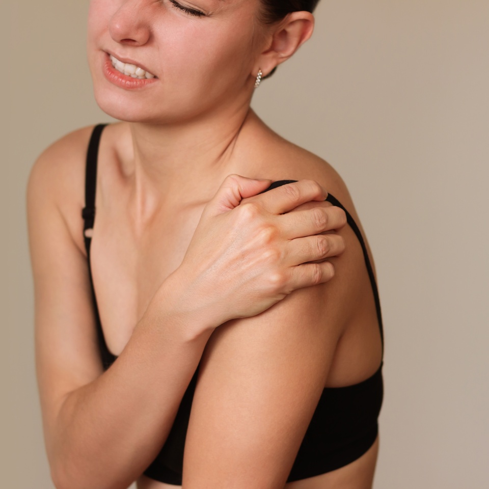 Young caucasian brunette woman in black underwear, holding her sore shoulder with her hand, suffering from shoulder pain, arthritis. Isolated on a beige background. Shoulder injury.