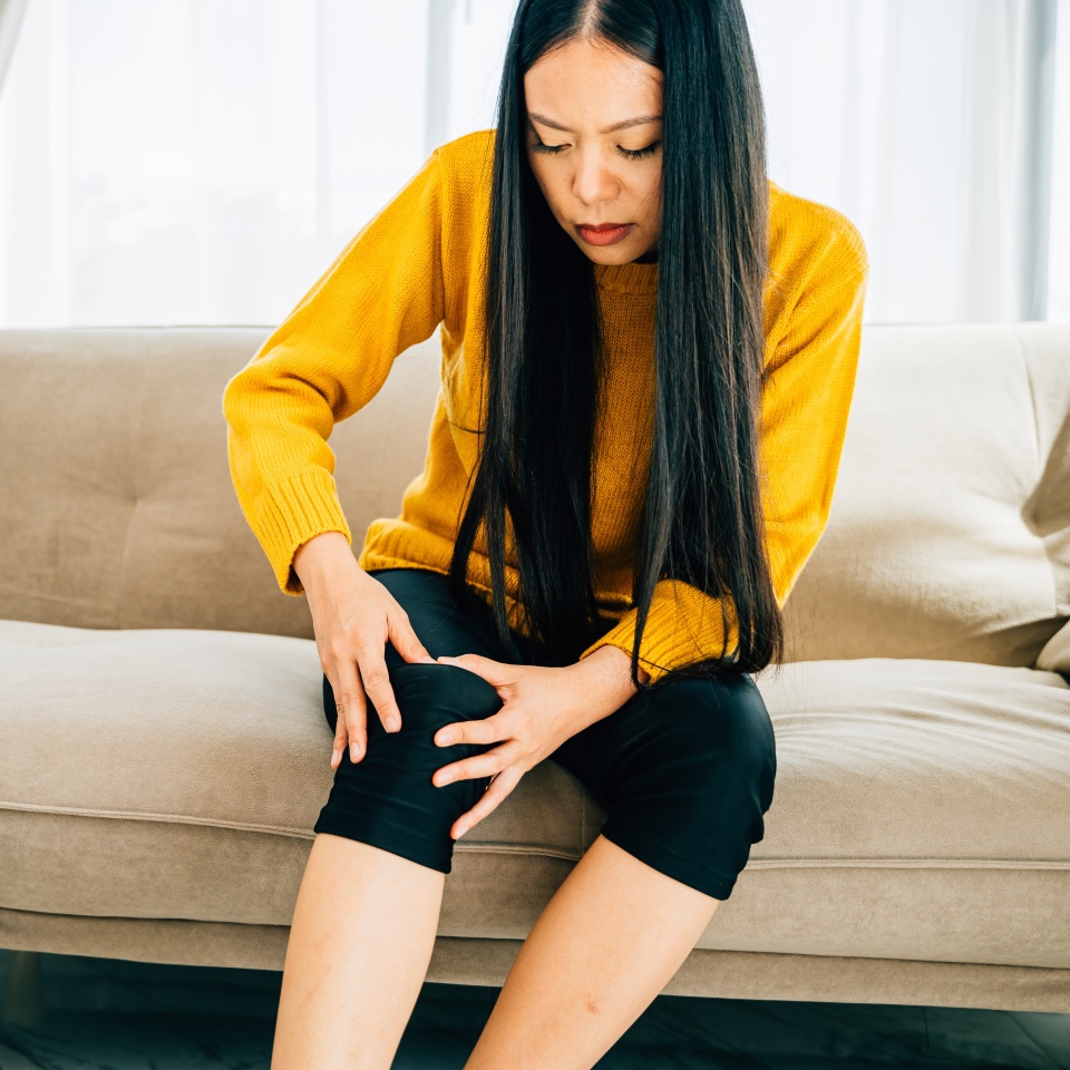 A woman seated on sofa holding a painful knee due to chronic tendon arthritis. Depicting Health Care and Medical Concept highlighting pain and tendon inflammation.
