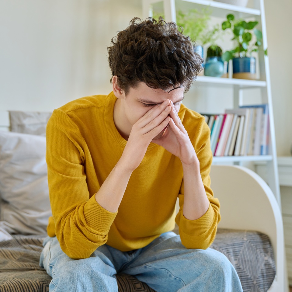 Sad upset young man sitting on couch at home, touching his head with hands