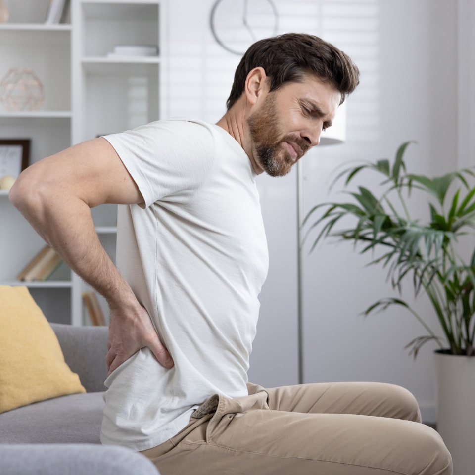 Man sitting on couch holding lower back, experiencing back pain, discomfort or injury. Concept of health issues, chronic pain, posture problems, or muscle strain.