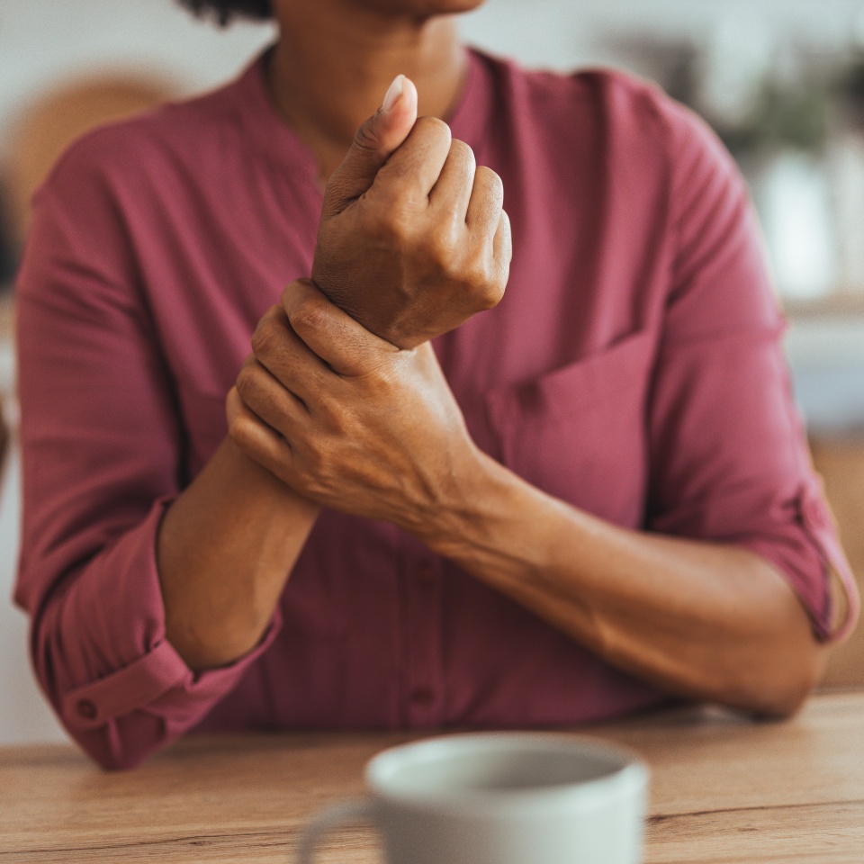 A woman in a pink shirt sits at a table, holding her wrist in discomfort, symbolizing wrist pain or arthritis in a home setting.