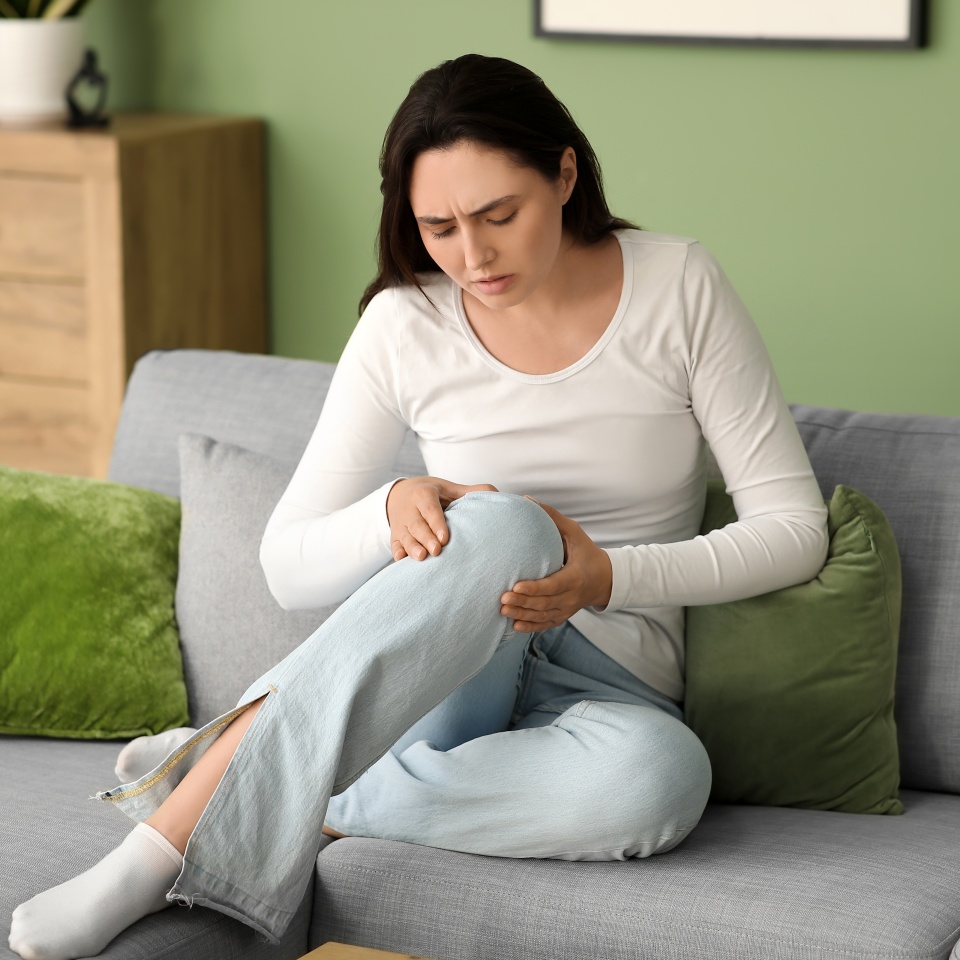 Young woman suffering from pain in knee on sofa at home
