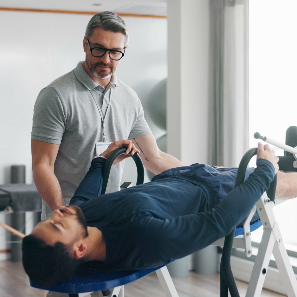 Physiotherapist, person and recovery on fitness bench for exercise, help or progress at clinic. People, biokinetics and support with guide for rehabilitation process with inversion table at hospital
