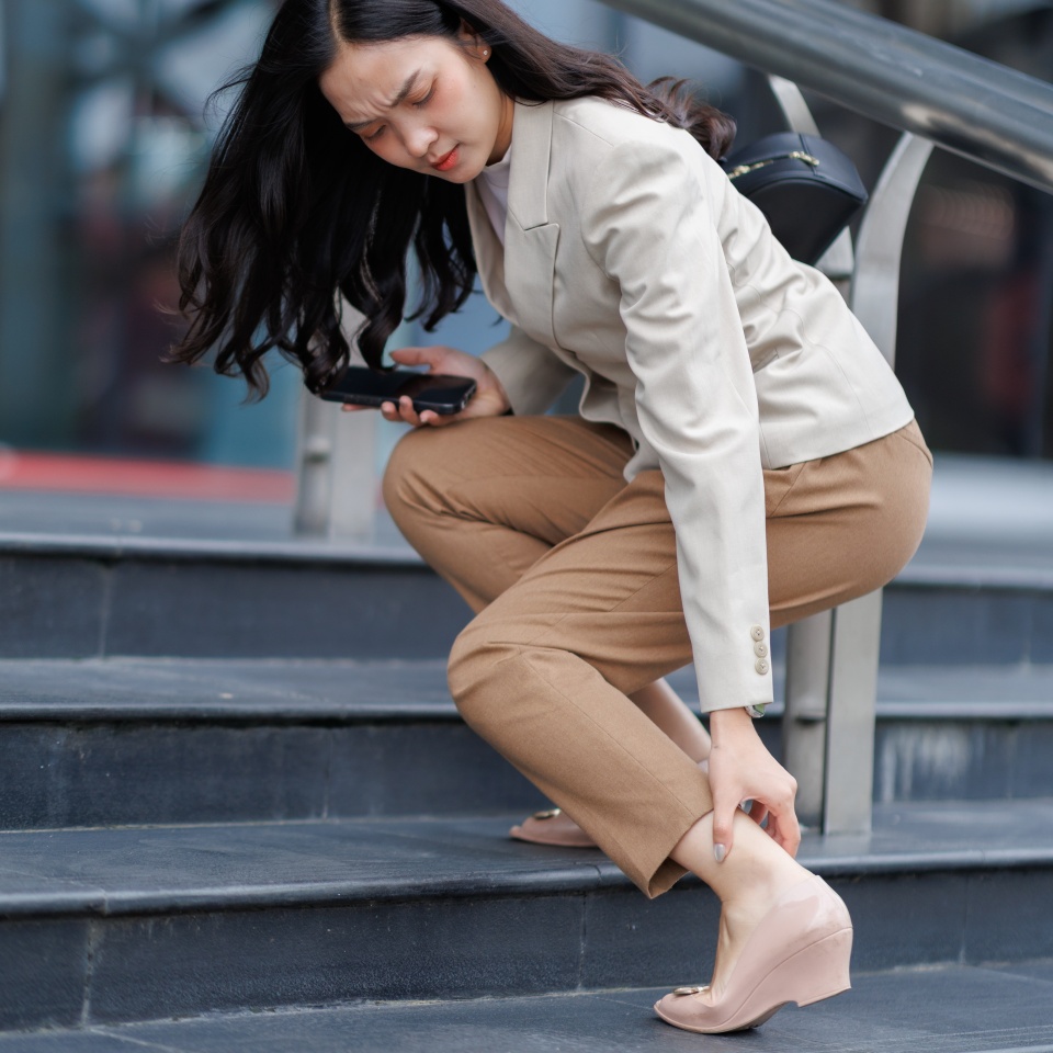 Young businesswoman holding her smartphone and touching her painful ankle while walking down the stairs