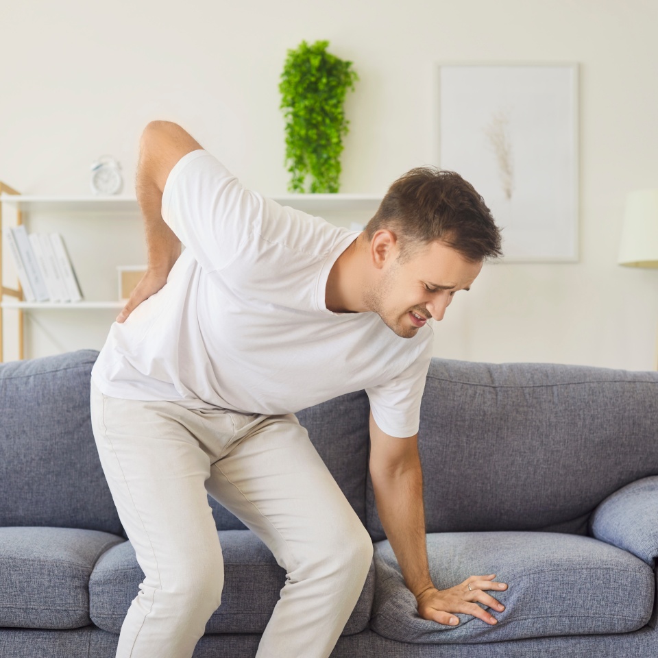 Sick man with lower back pain standing up from sofa in living room at home, holding lumbar with hand. Sad young person suffering from sciatica, pinched nerve or inflammation of intervertebral disc