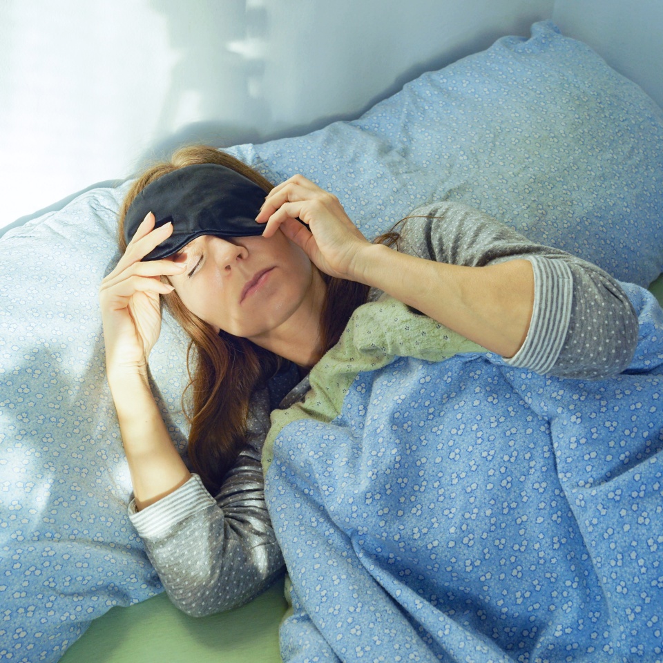 Woman adjusting eye mask while lying down during an acute migraine attack in a daylight bedroom. Represents light-triggered pain response, migraine self-care technique, and darkness therapy necessity.