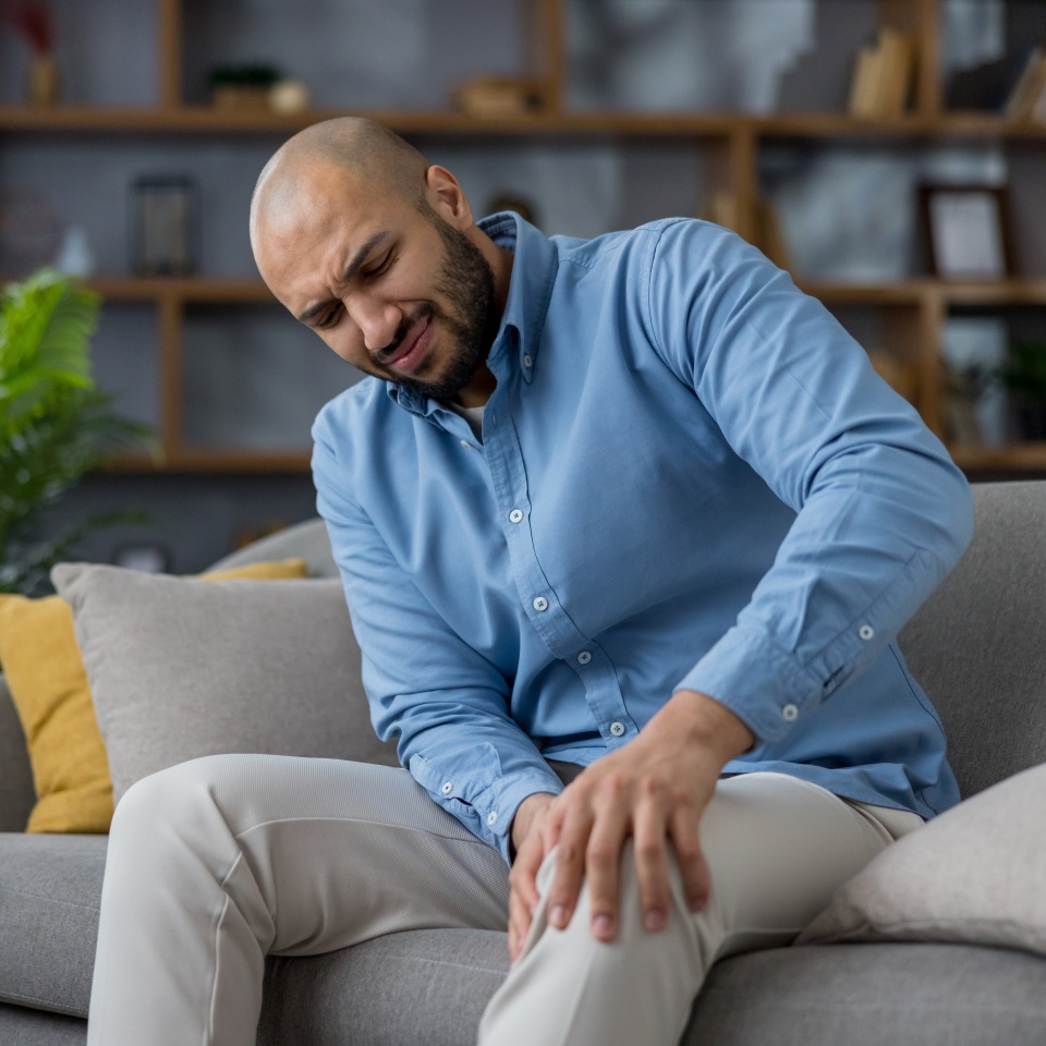 Man feeling intense pain in his knee, sitting on a gray sofa at home, touching his leg, experiencing discomfort and health issues, requiring care for joint and muscle problems