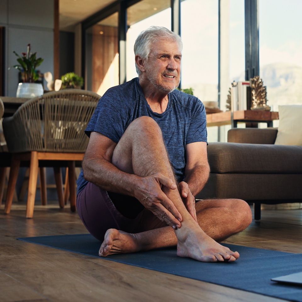 Fitness, laptop and senior man with ankle pain, injury or accident with virtual exercise or training. Computer, health and elderly male person with muscle sprain for online workout in living room.