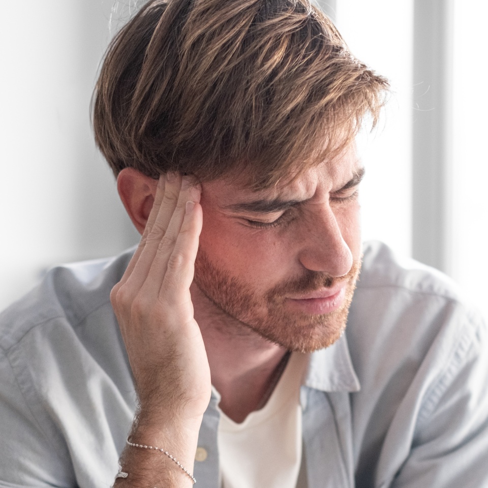 An attractive young man with a beard sits on the couch at home in a relaxed, modern setting, rubbing his temples to relieve a headache. A bright light comes in from the window.