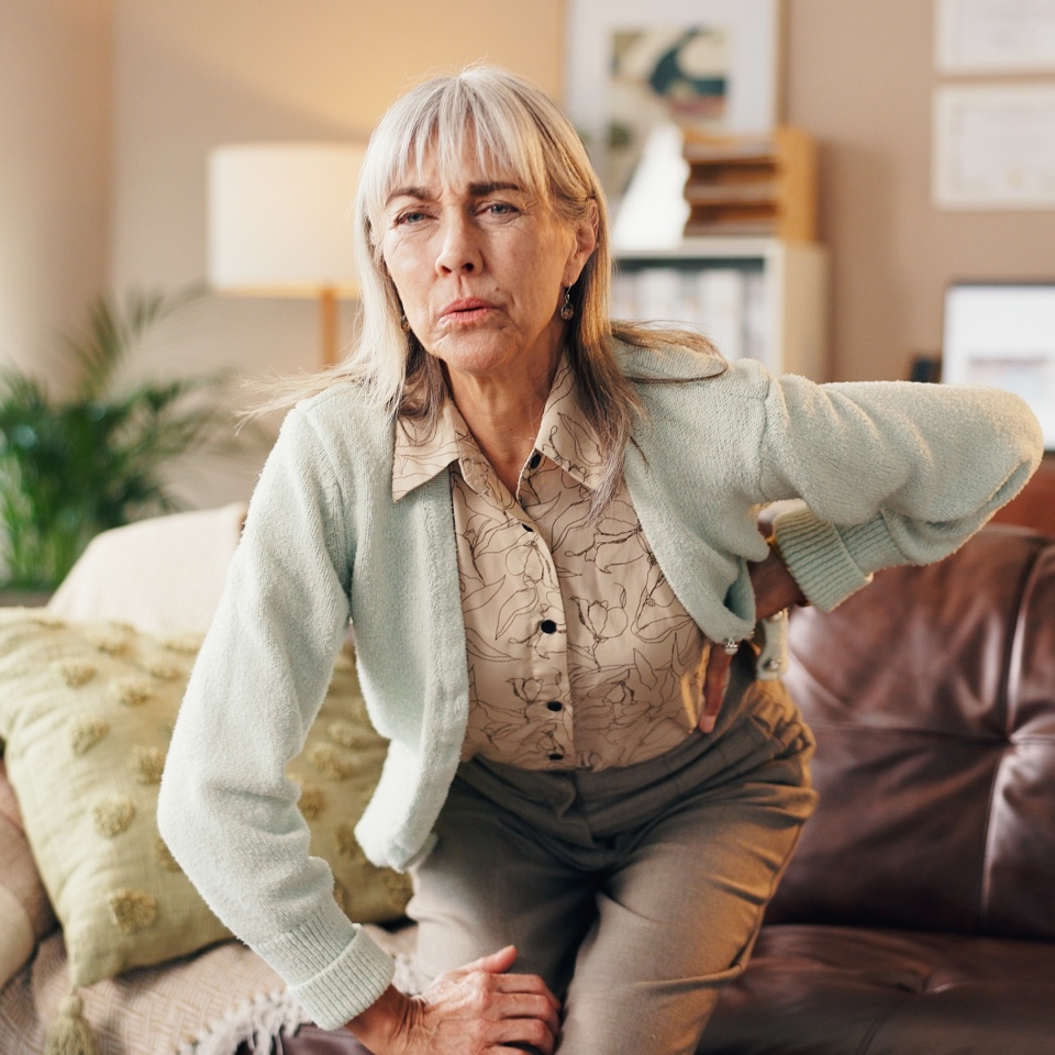 Back pain, posture and senior woman on sofa in living room of home for injury or osteoporosis. Discomfort, muscle fatigue and spinal stenosis with old person in apartment for arthritis symptoms