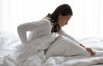 Side view of millennial woman woke up sitting in bed, unhealthy female woke up feels severe lower back pain during period, pinched nerve discomfort in vertebrae, herniated spinal disc symptoms concept