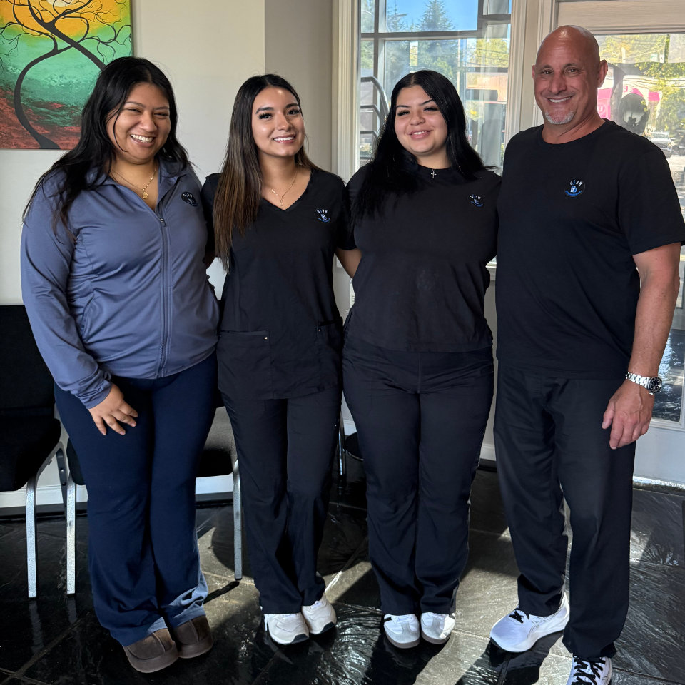 Four people standing together in an office, two wearing black scrubs, one wearing a black t-shirt, and one wearing a grey zip-up jacket.
